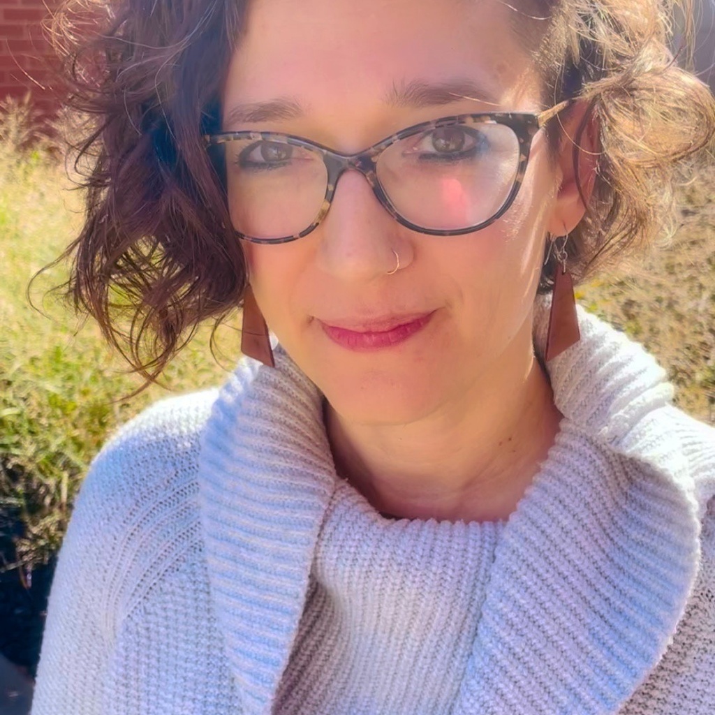 Natalie Fixmer-Oraiz in a grey sweater standing outside with glasses and brown triangle earrings